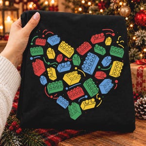 Puede incluir: Una camiseta negra con un estampado gráfico de bloques de construcción de plástico de colores dispuestos en forma de corazón. Los bloques son rojos, azules, amarillos y verdes, con pequeñas líneas curvas por todo el diseño.