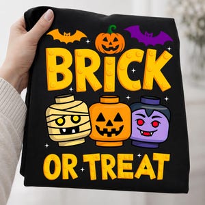 May include: A black t-shirt featuring a graphic with the text BRICK OR TREAT in yellow block letters. The design includes a jack-o'-lantern, an orange bat, a purple bat, and three toy-style heads: a mummy, a pumpkin, and a vampire.