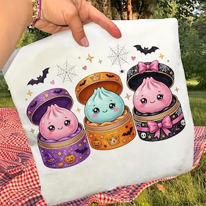 Puede incluir: Una bolsa de tela blanca con un estampado gráfico de tres adorables dumplings sonrientes en recipientes coloridos y decorados. El diseño incluye pequeñas calabazas, murciélagos, telarañas, estrellas y lazos rosas sobre un fondo blanco.
