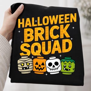 May include: A black t-shirt featuring the yellow text HALLOWEEN BRICK SQUAD in a blocky, studded font. Below the text are four cartoon character heads resembling toy building blocks: a mummy, a jack-o'-lantern, a skull, and a green monster.