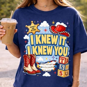 May include: A navy blue t-shirt featuring a graphic print with the text "I KNEW IT I KNEW YOU". The design includes a yellow sheriff star with "INFINITY AND BEYOND", a red cowboy hat, red and white cowboy boots, colorful beads, and alphabet blocks.