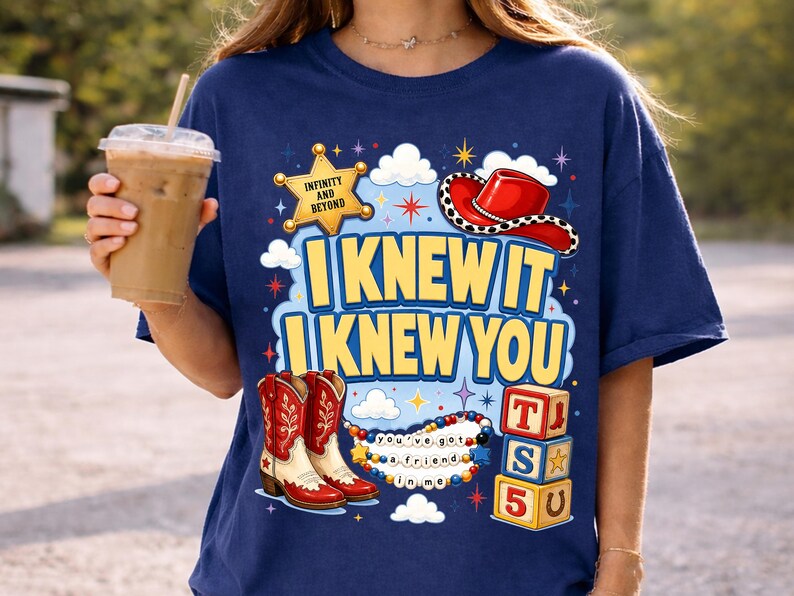 May include: A navy blue t-shirt featuring a graphic print with the text "I KNEW IT I KNEW YOU". The design includes a yellow sheriff star with "INFINITY AND BEYOND", a red cowboy hat, red and white cowboy boots, colorful beads, and alphabet blocks.