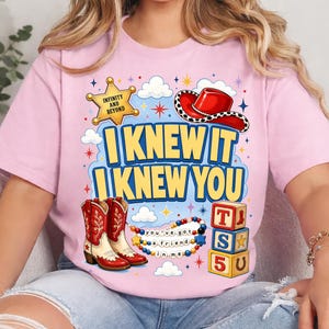 May include: A light pink t-shirt featuring a graphic print with the text "I KNEW IT I KNEW YOU". The design includes a yellow sheriff star, a red cowboy hat, red and white cowboy boots, a beaded necklace, and three stacked alphabet blocks.