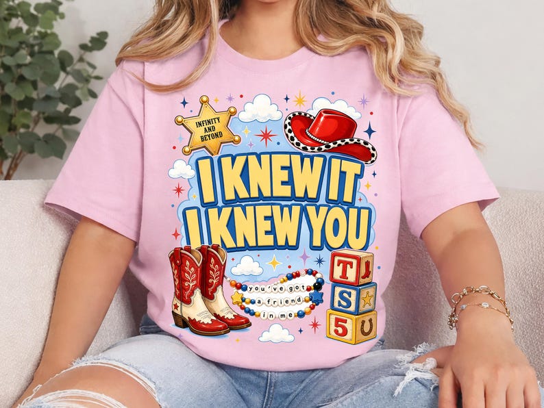 May include: A light pink t-shirt featuring a graphic print with the text "I KNEW IT I KNEW YOU". The design includes a yellow sheriff star, a red cowboy hat, red and white cowboy boots, a beaded necklace, and three stacked alphabet blocks.