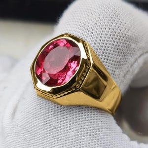 Könnte beinhalten: Ein goldener Ring mit einem großen, facettierten, oval geformten, roten Edelstein. Der Ring hat einen dicken Schaft und eine dekorative Lünette um den Stein. Auf weißem Hintergrund.