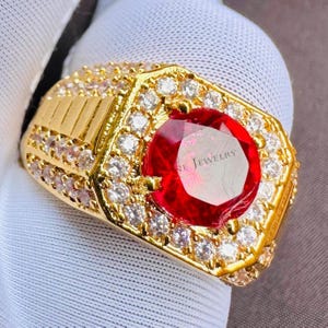 Könnte beinhalten: Ein goldener Ring mit einem großen, runden, roten Edelstein in der Mitte, umgeben von kleineren, klaren Edelsteinen. Der Ring hat eine rechteckige Oberfläche mit einem strukturierten Design an der Seite. Der Text "NE JEWELRY" ist auf dem Edelstein sichtbar.