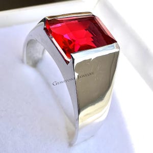 Könnte beinhalten: Ein silberner Ring mit einem großen, rechteckigen, facettierten roten Edelstein. Der Ring hat eine polierte Oberfläche und die Worte "GEMSTONE JEWELRY" sind auf der Seite sichtbar.