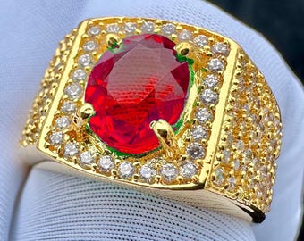 May include: A gold-colored ring featuring a large, oval-cut, red gemstone as the centerpiece. The ring is encrusted with small, clear gemstones around the central stone and along the band, creating a luxurious appearance.