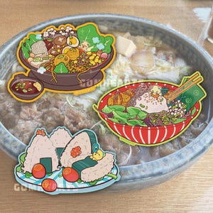 May include: Three die-cut stickers featuring illustrated food: a brown bowl of ramen with vegetables and mushrooms, a red bowl of noodles with a small yellow bear character, and a plate of three rice balls with salmon roe and a small bear.