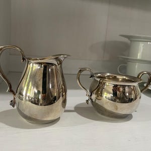 May include: Two silver-colored serving pieces, a creamer and a sugar bowl, sit on a white shelf. The creamer has a handle and a spout. The sugar bowl has two handles. Both pieces reflect the surrounding environment.
