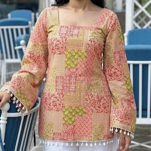 Puede incluir: Top con estampado de patchwork con mangas largas y cuello cuadrado. El top presenta una mezcla de patrones rojos, verdes y beige. Las mangas y el dobladillo están adornados con borlas blancas. Combinado con pantalones blancos.