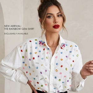 May include: A white shirt embellished with a variety of colourful gemstones. The long-sleeved shirt has a classic collar. Text on the left reads "NEW ARRIVAL: THE RAINBOW GEM SHIRT EXCLUSIVELY AVAILABLE".