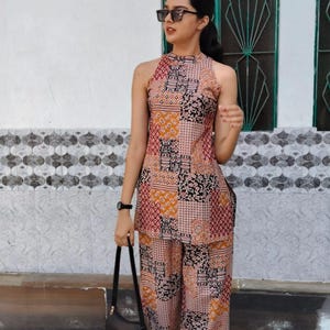 Puede incluir: Conjunto de top halter y pantalón de pernera ancha a juego. El conjunto presenta un diseño de patchwork en tonos rojos, naranjas, negros y crema. El atuendo se complementa con un bolso negro y gafas de sol.