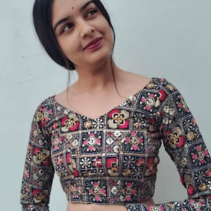 May include: A long-sleeved, embroidered blouse with a square pattern. The blouse features a V-neck and is adorned with gold, red, and pink floral designs on a dark blue background. The sleeves and neckline are also embellished with intricate embroidery.