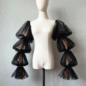 May include: Black tulle sleeves with multiple layers of fabric, displayed on a mannequin. The sleeves have a layered, puffy design, with each layer becoming wider towards the end. The fabric is sheer, allowing a glimpse of the mannequin's arm.