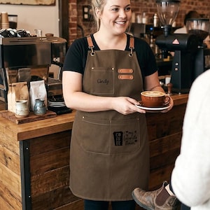 May include: A brown apron with leather accents and the name "Cindy" embroidered on the front. The apron has two pockets and a coffee-themed graphic. The barista is holding a cup of coffee with latte art.