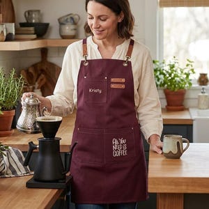 May include: A burgundy apron with leather accents and the text "All You Need Is Coffee." The apron has two pockets and the name "Kristy" embroidered on the chest. A coffee maker and mug are also visible.