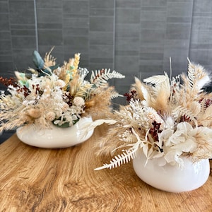 Peut inclure: Deux vases en céramique blanche remplis de fleurs et de graminées séchées. Les compositions présentent un mélange d'éléments beiges, crème et bordeaux, créant une esthétique naturelle et texturée. Les vases sont posés sur une surface en bois.