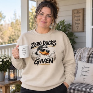 May include: A beige crewneck sweatshirt featuring a graphic of a duck wearing a backwards baseball cap and sunglasses, making hand gestures. The text "ZERO DUCKS GIVEN" is printed in black above and below the duck. A white mug with "Good Things Today" is held.
