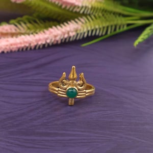 Pode incluir: Um anel dourado com um design de tridente e uma pedra preciosa verde. O anel está sobre um fundo roxo com folhagem rosa e verde. Uma peça de joalharia única.