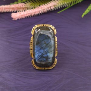May include: A rectangular ring featuring a large, iridescent labradorite stone. The stone displays blue and gray hues, set in a gold-toned metal frame with a decorative edge. The ring is a statement piece, suitable for adding a touch of elegance.