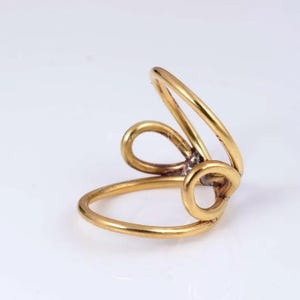 Puede incluir: Un anillo dorado con un diseño abierto y único. El anillo presenta una forma de bucle con un elemento circular y líneas curvas. La joya está sobre un fondo blanco, resaltando sus intrincados detalles.