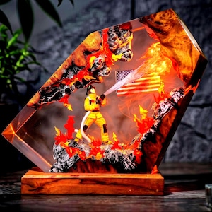 Puede incluir: Una lámpara decorativa de resina y madera que muestra a un bombero con equipo amarillo sosteniendo una manguera, rociando agua hacia una bandera estadounidense brillante. La escena está encerrada en un bloque de resina angular y transparente.