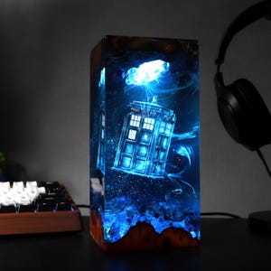 May include: A rectangular resin lamp featuring a blue glowing image of a police box, with the text "POLICE BOX" visible. The lamp has a wooden base and is surrounded by blue light effects. A keyboard and headphones are in the background.