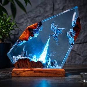 May include: A trapezoidal resin and wood sculpture featuring a miniature dragon breathing a bright blue light onto a castle landscape. The piece is set on a rectangular wooden base, combining clear resin with natural wood grain and blue illumination.