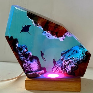 May include: A decorative lamp featuring a marine scene encased in resin. The lamp has a wooden base and a geometric shape. The scene includes a manta ray, divers, and underwater rock formations, illuminated with blue and pink LED lights.