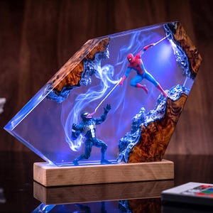 Puede incluir: Una lámpara decorativa de resina y madera con una figura de Spider-Man en traje rojo y azul y una figura de Venom en traje negro. Las figuras están encerradas en un bloque de resina hexagonal transparente con iluminación LED azul y humo.