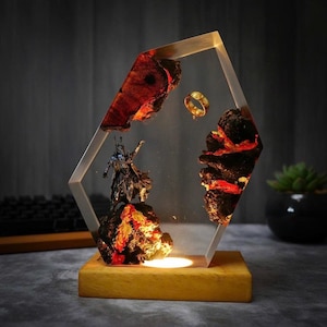 May include: A decorative resin lamp featuring a fantasy scene with a miniature figure, a gold ring, and fiery rock formations. The lamp has a wooden base and a clear, geometric frame, with a warm light illuminating the scene.