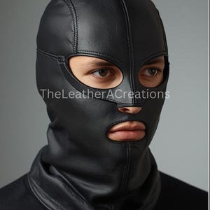 May include: A black leather hood with eye and mouth openings. The hood covers the head and neck, with visible stitching details. The text "TheLeatherACreations" is printed on the lower left side of the hood.