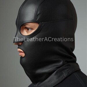 May include: Black leather hood with eye and mouth openings. The hood is stitched and covers the head and neck. The text "TheLeatherACreations" is visible on the lower left side of the hood.