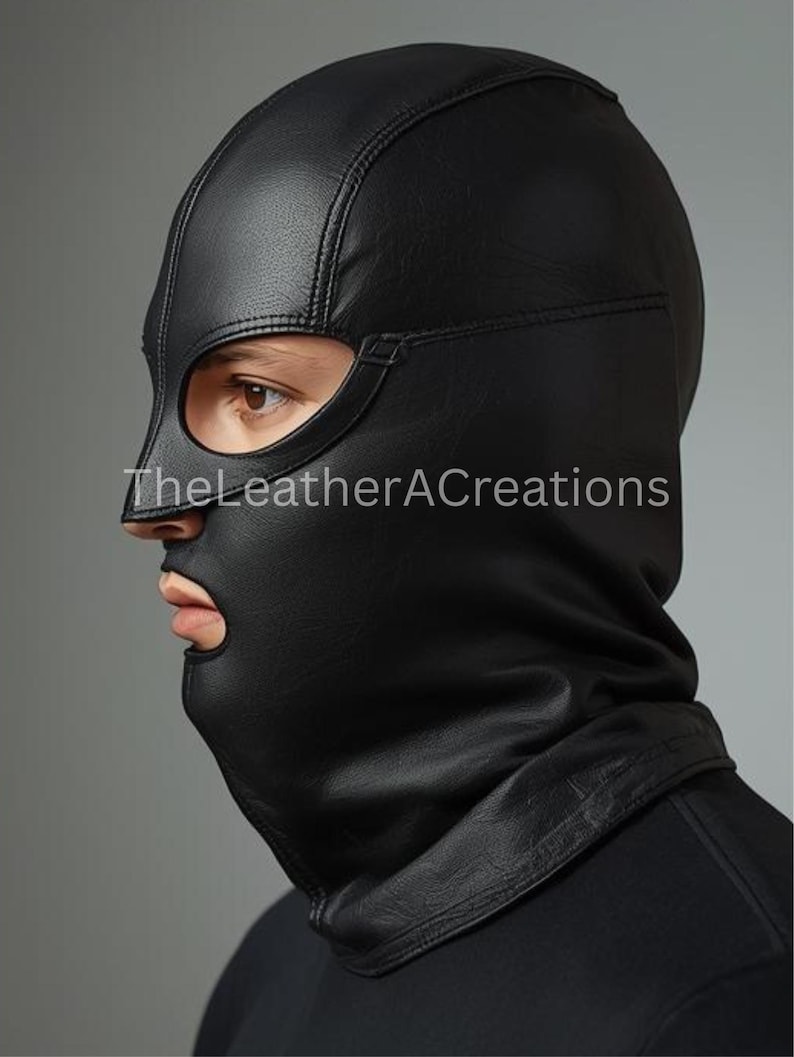 May include: Black leather hood with eye and mouth openings. The hood is stitched and covers the head and neck. The text "TheLeatherACreations" is visible on the lower left side of the hood.