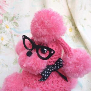 Puede incluir: Un juguete de peluche de caniche rosa con gafas negras y una pajarita negra con lunares blancos. El caniche tiene una textura esponjosa y está sobre un fondo floral.