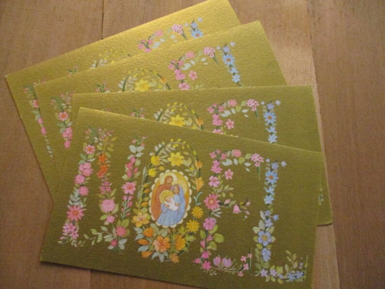 Puede incluir: Cuatro tarjetas de felicitaci&oacute;n doradas con dise&ntilde;os florales y una imagen de la Sagrada Familia. Las tarjetas presentan flores coloridas en rosa, azul, amarillo y naranja. Las tarjetas est&aacute;n dispuestas sobre una superficie de madera.
