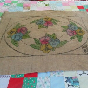 K&ouml;nnte beinhalten: Beige Jute-Textil mit floralem Design. Das Design zeigt eine kreisf&ouml;rmige Anordnung farbenfroher Stiefm&uuml;tterchen in Rosa, Gelb und Blau, mit gr&uuml;nen Bl&auml;ttern. Das Textil hat eine schwarze Kontur und ein gestempeltes Logo.