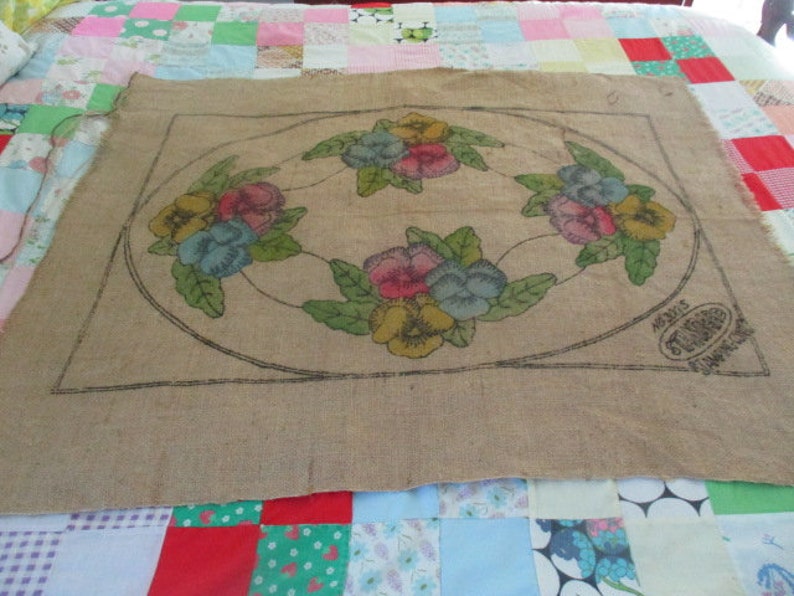 K&ouml;nnte beinhalten: Beige Jute-Textil mit floralem Design. Das Design zeigt eine kreisf&ouml;rmige Anordnung farbenfroher Stiefm&uuml;tterchen in Rosa, Gelb und Blau, mit gr&uuml;nen Bl&auml;ttern. Das Textil hat eine schwarze Kontur und ein gestempeltes Logo.