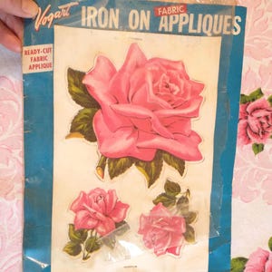 May include: A package of Vogart iron-on fabric appliqués featuring pink rose designs. The package includes a large rose and two smaller roses with green leaves. Text on the package reads "Iron On Appliques" and "Easy to Iron On Washable."