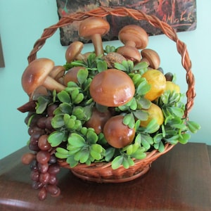 May include: A woven brown wicker basket filled with artificial green foliage, smooth brown mushroom figures, yellow fruit, and a cluster of dark red grapes. The basket features a curved handle and is arranged with various decorative elements.