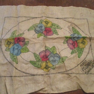 K&ouml;nnte beinhalten: Ein rechteckiges St&uuml;ck Jute mit einem floralen Design. Das Design umfasst ein zentrales Oval mit Clustern farbiger Stiefm&uuml;tterchen in Rot, Blau und Gelb, umgeben von gr&uuml;nen Bl&auml;ttern. Die Kanten sind unfertig.