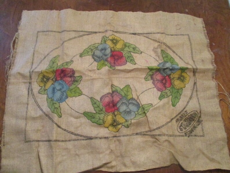 K&ouml;nnte beinhalten: Ein rechteckiges St&uuml;ck Jute mit einem floralen Design. Das Design umfasst ein zentrales Oval mit Clustern farbiger Stiefm&uuml;tterchen in Rot, Blau und Gelb, umgeben von gr&uuml;nen Bl&auml;ttern. Die Kanten sind unfertig.