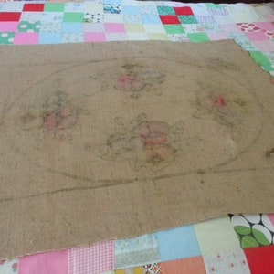 K&ouml;nnte beinhalten: Ein St&uuml;ck Jute-Stoff mit einem Blumendesign und einer ovalen Kontur. Das Blumendesign zeigt rosa und gr&uuml;ne Blumen. Der Stoff liegt auf einem bunten Patchwork-Quilt.
