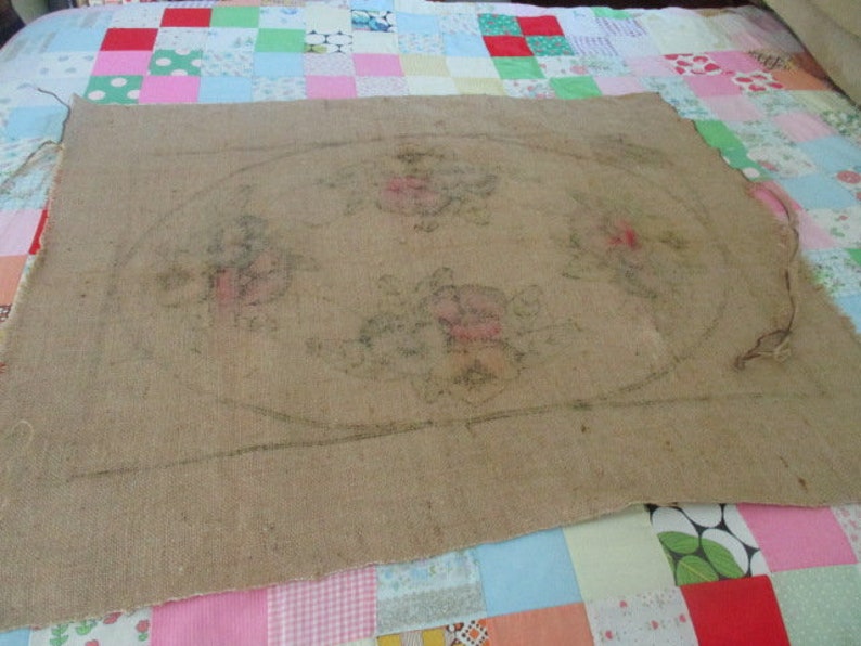 K&ouml;nnte beinhalten: Ein St&uuml;ck Jute-Stoff mit einem Blumendesign und einer ovalen Kontur. Das Blumendesign zeigt rosa und gr&uuml;ne Blumen. Der Stoff liegt auf einem bunten Patchwork-Quilt.