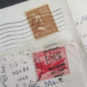 Op de afbeelding: Close-up van vintage enveloppen met postzegels. E&eacute;n postzegel is bruin met een portret en leest "1 1/2 CENTS 12". Een andere is rood met een vliegtuigafbeelding en "AIR MAIL". De enveloppen hebben handgeschreven notities en poststempels.