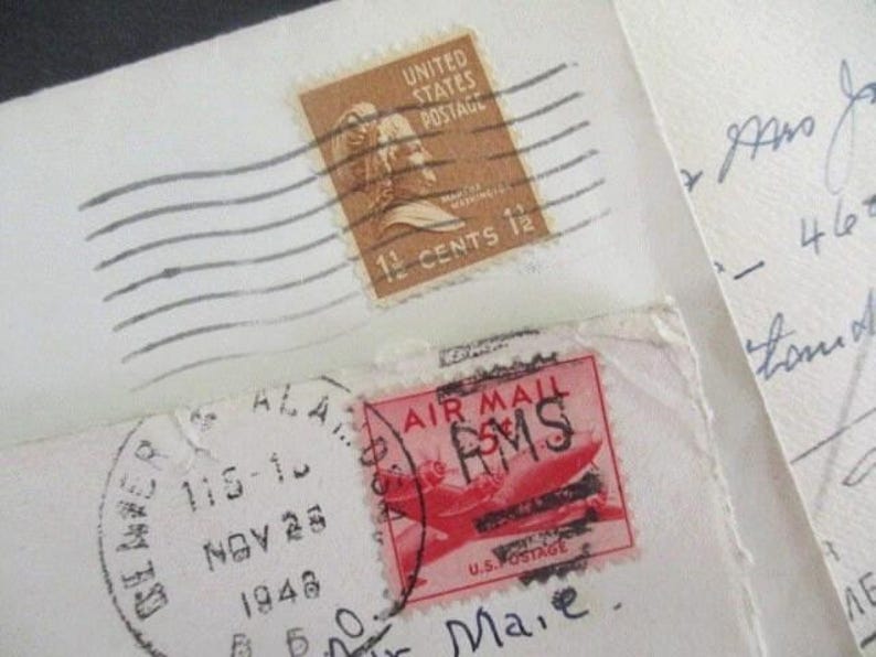 Op de afbeelding: Close-up van vintage enveloppen met postzegels. E&eacute;n postzegel is bruin met een portret en leest "1 1/2 CENTS 12". Een andere is rood met een vliegtuigafbeelding en "AIR MAIL". De enveloppen hebben handgeschreven notities en poststempels.