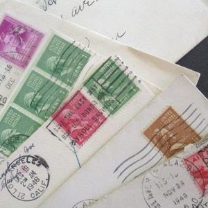 Op de afbeelding: Close-up van vintage enveloppen met postzegels. De postzegels zijn in verschillende kleuren, waaronder groen, paars en bruin. Sommige enveloppen hebben poststempels uit Denver en Los Angeles, daterend uit 1948.