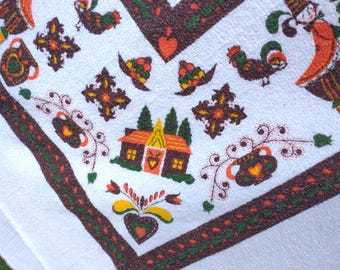 Könnte beinhalten: Ein weißes Textilgewebe mit braunem Rand und einem wiederkehrenden Muster aus bunten Stickmotiven, darunter ein kleines Haus mit orangefarbenem Dach, Hähne, florale Herzformen und dekorative Kreuzmuster in Grün-, Orange- und Brauntönen.