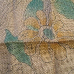 May include: Close-up of a burlap fabric with a floral design. The design features a large flower with yellow petals, a blue center, and teal leaves. The fabric has a rough texture and visible weave. Numbers are visible on the design.